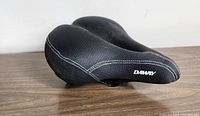 Side-angle view of black textured padded Daway C99 bike saddle with white stitching on wooden surface.