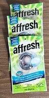 Three packages of Affresh washer cleaner tablets stacked and leaning against each other on a wooden surface. Each package is green and white with the Affresh logo and product information visible.