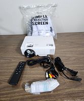 Image showing white mini projector, black remote, power cord, AV cable, projection screen in plastic, and sealed accessory pack on a wooden surface.