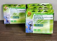 Photo showing six Duck brand window kits in green packaging, some with 10 pieces and some with 5 pieces of crystal clear shrink film for window insulation