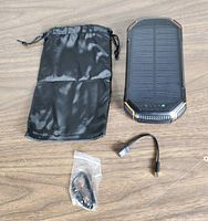 Black solar power bank with visible solar panel and green LED light, showing textured edges and rugged design along with black carrying pouch and two USB cables.