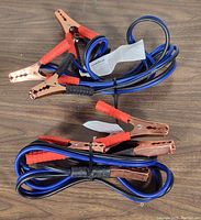 Two sets of new Goodyear booster cables coiled on a wooden surface. Each cable features copper clamps with red and black insulated grips and blue cables tied with plastic ties.