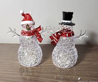 Two clear LED snowmen figures on a wooden surface, one with a red Santa hat and one with a black top hat, both with red scarves and silver twig arms.