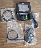 Ingenico iSC350 electronic payment terminal with power adapter and cables all sealed in plastic bags on a wooden surface.