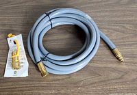 Photo of coiled grey outdoor gas hose with brass fittings and attached product tag on wooden surface.