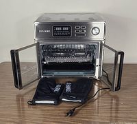Stainless steel air fryer oven with dual glass doors open, showing cooking basket, rack, crumb tray and two black oven mitts on wood surface