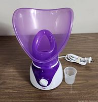 Photo of facial steamer unit with purple face mask attachment, white base, control dial, and power cord with measuring cup included.