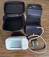 Top-view photo showing the Omron BP5350 digital blood pressure monitor device with an LCD screen, three buttons, attached grey tube leading to a black arm cuff, four AA batteries beside the device, and an open black carry case.
