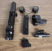 Pinty 3-9x32EG rifle scope and accessories spread out on wood surface, showing scope, laser sight, lens covers, and mounting rails.