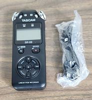 Tascam DR-05 handheld digital audio recorder placed on wood surface, showing front side with control buttons and screen, beside sealed plastic bag containing cables or accessories.