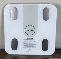 Top view of True Know Intelligent Body Fat Scale showing white surface with metal sensors, central circular logo, and caution label.