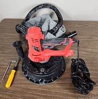 Handheld red and black drywall sander with attached dust collection bag, power cord coiled beside it, detachable handle, and hex key tool.