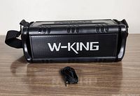 Black W-King D8 portable wireless speaker placed on a wooden surface with shoulder strap attached. A black auxiliary cable is coiled and placed in front.