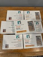 Seven sealed boxes of Retekess V112 FM/AM portable radios stacked on a wooden surface. Each box shows the radio image and model details.