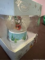 Side view of Disney snow globe in original box, showing Mickey and Minnie inside and part of blue base with Christmas theme.