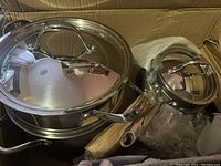 Photo showing stainless steel pots and pans stacked inside cardboard box, with lids and handles visible.