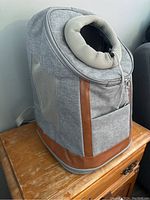 Light grey pet carrier backpack with brown trim and mesh ventilation on the sides and top circular mesh opening, sitting on wooden surface.