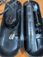 Opened black plastic travel case with Philips Sonicare electric toothbrush handle, two brush heads, and charging cord with US plug.