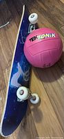 Photo showing galaxy-themed skateboard with white wheels and pink Tektonik volleyball side by side on wood floor.
