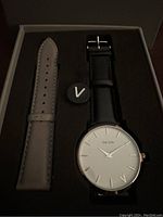 Watch in box with black leather strap and beige replacement strap, showing face of watch with white dial and minimalistic markers.