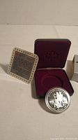Close view of the commemorative coin, certificate insert with text, and red velvet-lined presentation case.