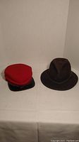 Two hats on white background: one red with black visor the other dark brown felt with braided band and feather.