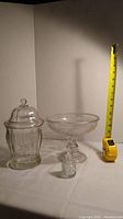 Three clear glass items: lidded candy dish, pedestal compote, small bottle with stopper. Tape measure for scale approximately 8 inches tall for compote.
