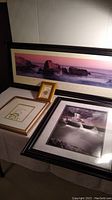 Four framed artworks grouped together on a surface including a panoramic sunset or sunrise landscape, black and white seascape, small gold frame artwork, and a matted botanical print.