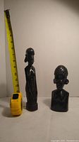 Two African ebony sculptures positioned next to a measuring tape showing heights around 9-11 inches, displayed against a plain background.
