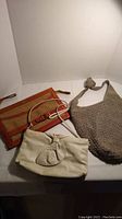 Three assorted vintage style purses arranged on a white surface; beige woven shoulder bag, cream leather clutch with bow, orange and brown striped clutch