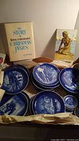 Photo of assorted Royal Copenhagen Christmas plates in blue and white with winter scenes, showing two hardcover books in the background.
