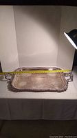Rectangular silver plated service platter laid flat showing length measurement with tape measure. Ornate edges and handles visible.