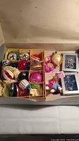 Box containing assorted vintage Christmas glass and metal ornaments with various colors and shapes including balls, teardrops, and character figures.
