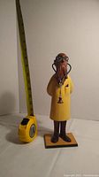 Wooden doctor figurine with yellow coat, glasses, and stethoscope shown next to tape measure for size reference.