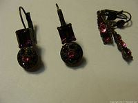 Close-up image of one pair of drop earrings featuring rectangular and round purple Swarovski crystals on black metal setting plus a partial view of another pair.