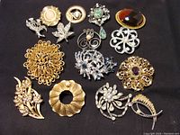 15 assorted vintage brooches laid flat on black fabric showing various designs including floral, leaf, swirl, and abstract motifs in gold-tone and silver-tone metals with stones and pearls.