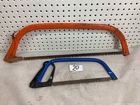 Photo showing the 24-inch orange frame bow saw and 12-inch blue frame bow saw side by side on a beige surface against a pegboard.