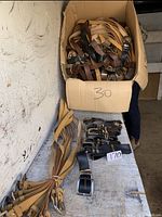 Box and table displaying large quantity of belts mainly in tan, black, and brown colors, packed loosely or bundled.