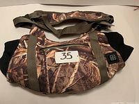 Camouflage heated hand warmer pouch laid flat showing front with zippered pocket, adjustable strap, and power button with label '35'.