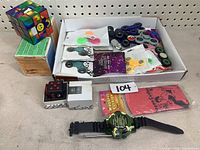 Box containing numerous plastic fidget spinners of various colors, small cube puzzles, a kids digital watch, and a whoops cushion.