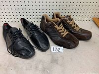 Pair of men's black leather dress shoes and brown casual sneakers placed side by side on the floor against a pegboard backdrop, showing sides and tops of shoes.