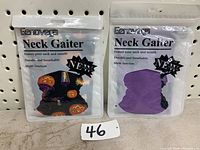 Two Genovega neck gaiters in resealable packaging hung on pegboard, one Halloween themed and one solid purple