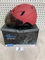 Red Odoland ski helmet displayed on its box showing front, side views with integrated large tinted goggle lens and ventilation holes.