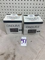 Two unopened boxes of ONDURA roofing screws and washers placed side by side on a gray surface with a pegboard background.