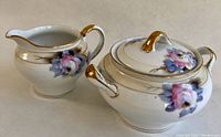Porcelain creamer and lidded sugar bowl set with multicoloured floral decoration and gold trim, shown from an angled side view