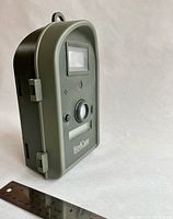 Closed view of green and black BirdCam camera with hanging loop on top, placed upright next to a ruler for size reference about 8.5 inches tall.