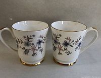 Photo of two white bone china tea mugs with gold trim and floral design, side by side showing overall appearance.