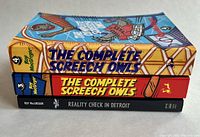 Side view showing the spines of three paperback books: two titled 'The Complete Screech Owls' volumes 3 and 4, and one titled 'Reality Check in Detroit' by Roy MacGregor.