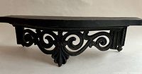 Angled view of black ornamental wall shelf showing edge profile and curved scroll design below shelf.