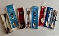 Seven vintage souvenir spoons from 1970s-1980s laid out side by side, showing packaging, various designs and tarnish.